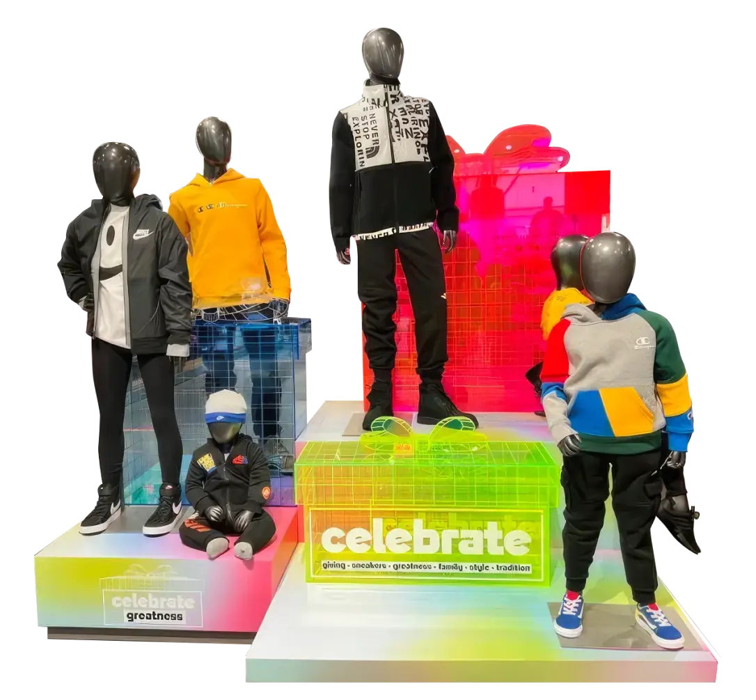 A display of five mannequins dressed in colorful, casual clothing. The mannequins are arranged on a multi-level platform with neon-colored accents. The outfits include hoodies, jackets, and pants in various colors such as black, yellow, blue, and gray. The background features vibrant neon lights in pink, green, and yellow hues with the words 'celebrate greatness' and 'celebrate' displayed on the platform.