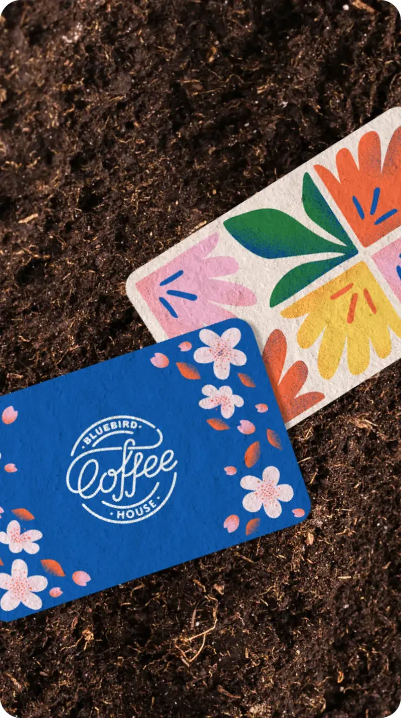 Three colorful cards on a soil background. The left card is blue with white flowers and the text 'Bluebird Coffee House' in white. The middle card has a floral design with pink, orange, yellow, and green colors. The right card is green with white leaves and the text 'Bluebird Coffee House' in white.